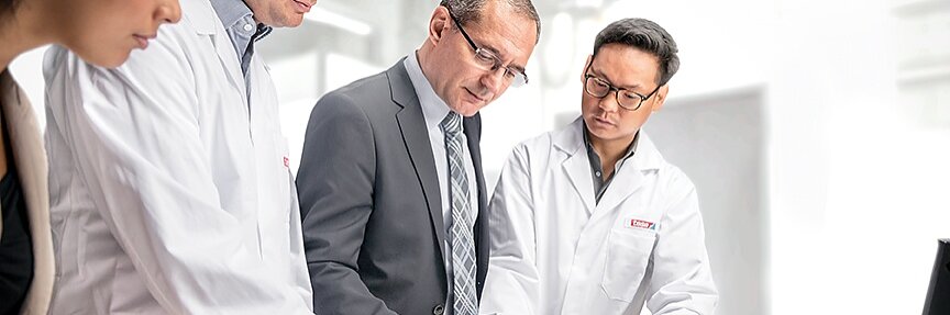 Four individuals are gathered around a table in a bright, modern workspace. Three are in white lab coats, and one is in a suit. They are engaged in reviewing documents and appear focused on their discussion about tesa tape. A laptop is partly visible on the table. (This text has been generated by AI)