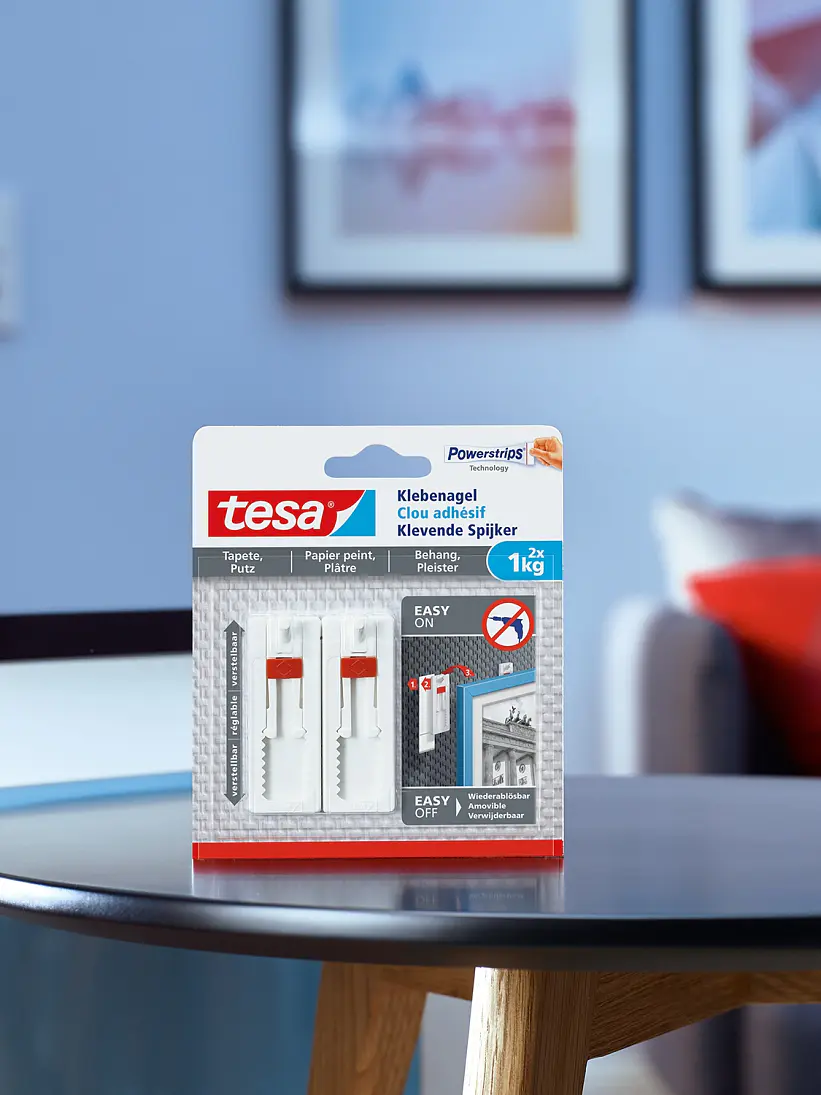 A pack of tesa tape adhesive nails for textured surfaces is displayed on a round black table in a modern living room setting. The product packaging is predominantly white and red, with blue and black elements. Blurred art and a door are visible in the background. (This text has been generated by AI)