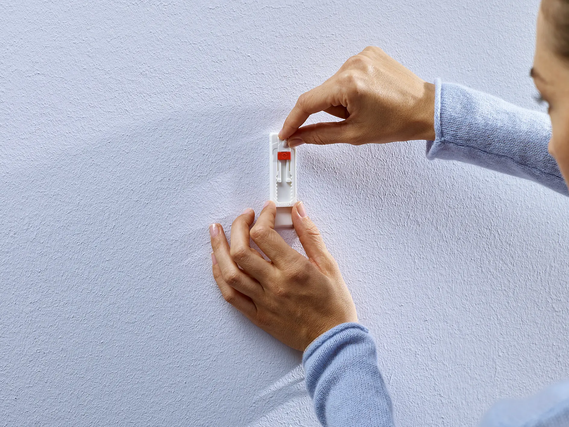 Hands of a person setting up a spring-loaded picture hook on a white wall. The individual is adjusting the hook, which features an orange tab and is designed for hanging items securely with tesa tape. The wall has a textured finish, and the persons sleeves are light blue. (This text has been generated by AI)