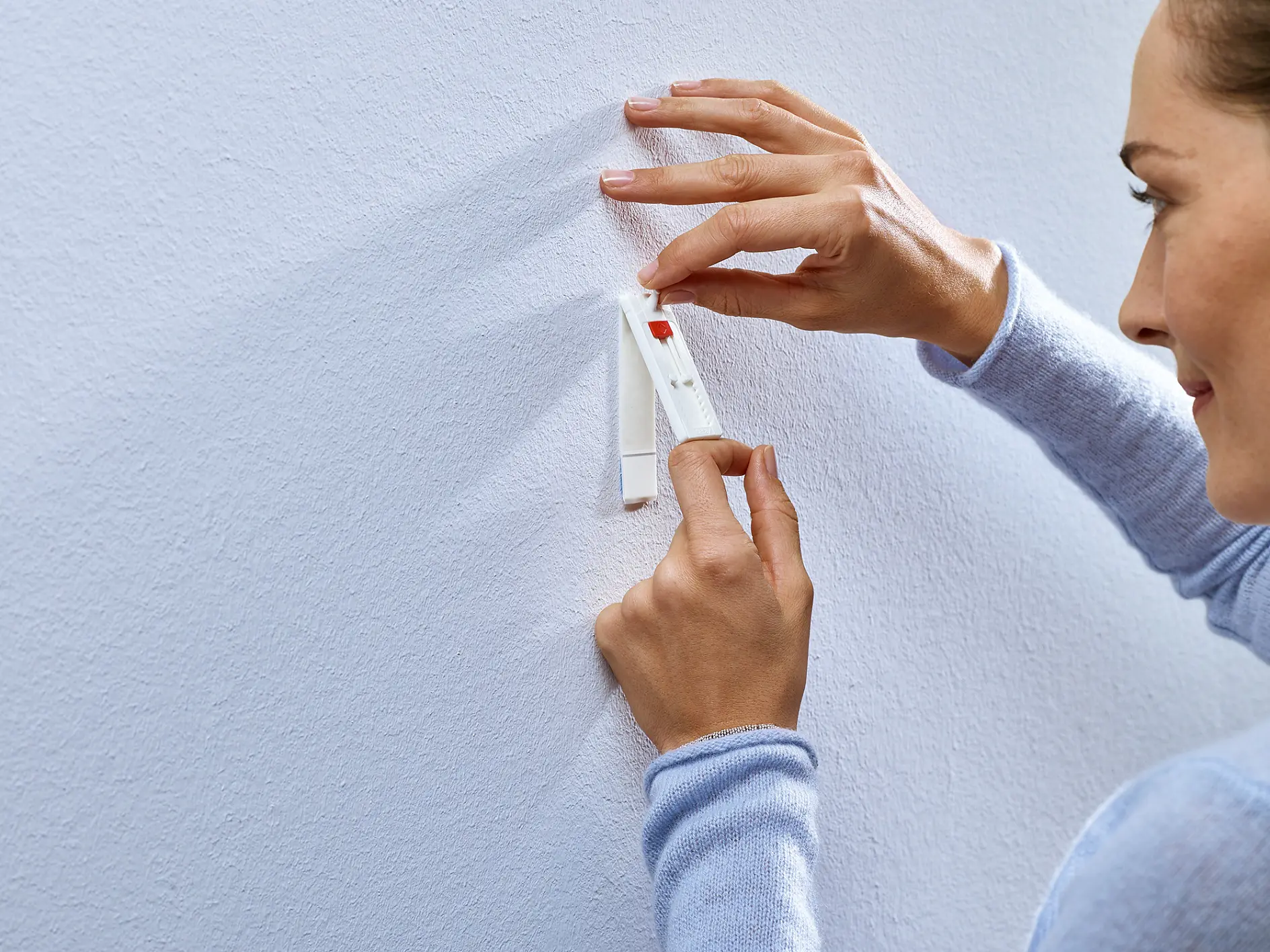 A person in a light blue sweater is attaching a white tesa tape strip to a white wall. They are peeling off the backing of the tesa tape while holding it in place. The image focuses on their hands and the wall. (This text has been generated by AI)