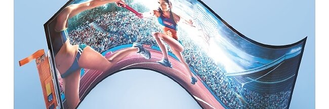 A flexible, curved display shows an image of two athletes on a running track engaging in a relay race. One athlete is passing the baton to another. The display is set against a light blue background, held securely with tesa tape for optimal visibility. (This text has been generated by AI)