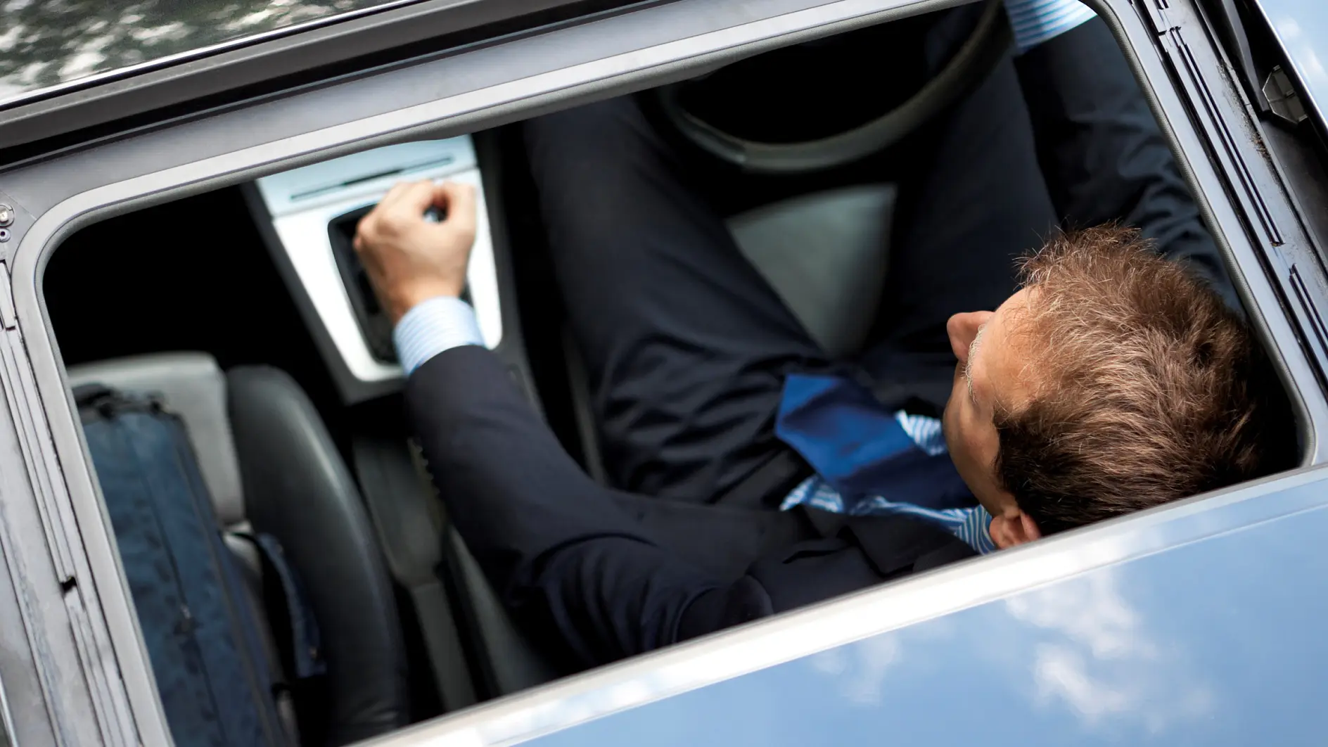 A man in a suit is seated inside a car, viewed from above through an open sunroof. He is adjusting a control on the cars dashboard. The interior appears contemporary, and a steering wheel is visible.