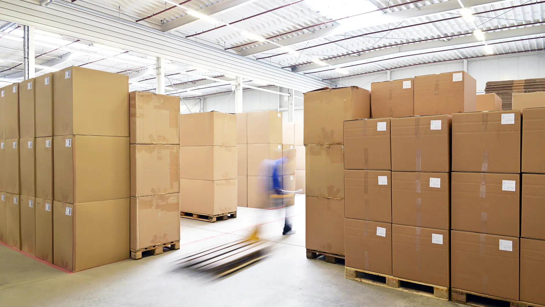 A large warehouse with stacks of cardboard boxes on wooden pallets. A person using a hand pallet truck is moving between the rows. The space is well-lit with natural light coming through the ceiling. The floor is concrete, and tesa tape is used throughout for securing packages and ensuring everything stays in place during transport.