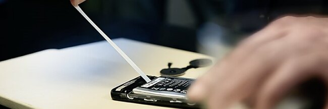 A persons hands are disassembling a smartphone using a tool. The back cover is removed, revealing the battery inside, secured with tesa tape. The scene suggests a repair or maintenance process on an electronic device. (This text has been generated by AI)