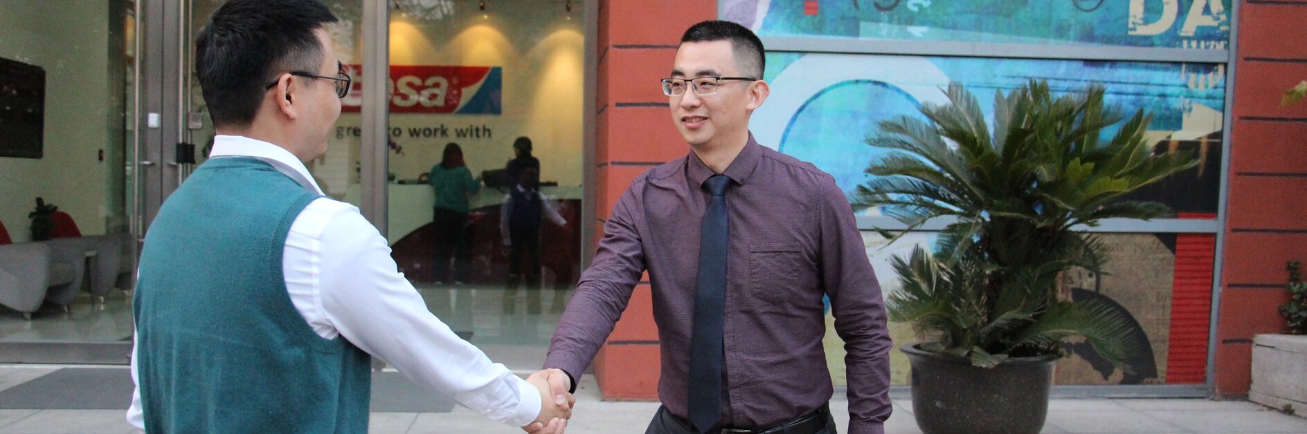 Two men are shaking hands in front of a building entrance. The man on the left is wearing a white shirt and teal vest, and the man on the right is wearing a purple shirt with a tie. Behind them is glass with artwork and the word tesa visible. (This text has been generated by AI)
