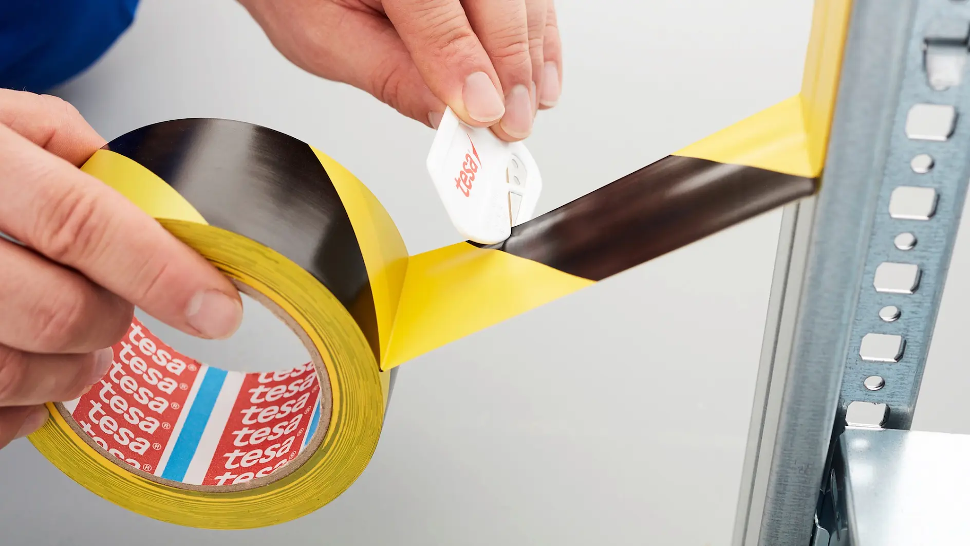 Close-up of hands cutting a piece of yellow and black striped tesa tape with a white cutting tool. The tesa tape is partially unrolled and attached to a metal surface. The inside of the tape roll shows the brand name tesa.