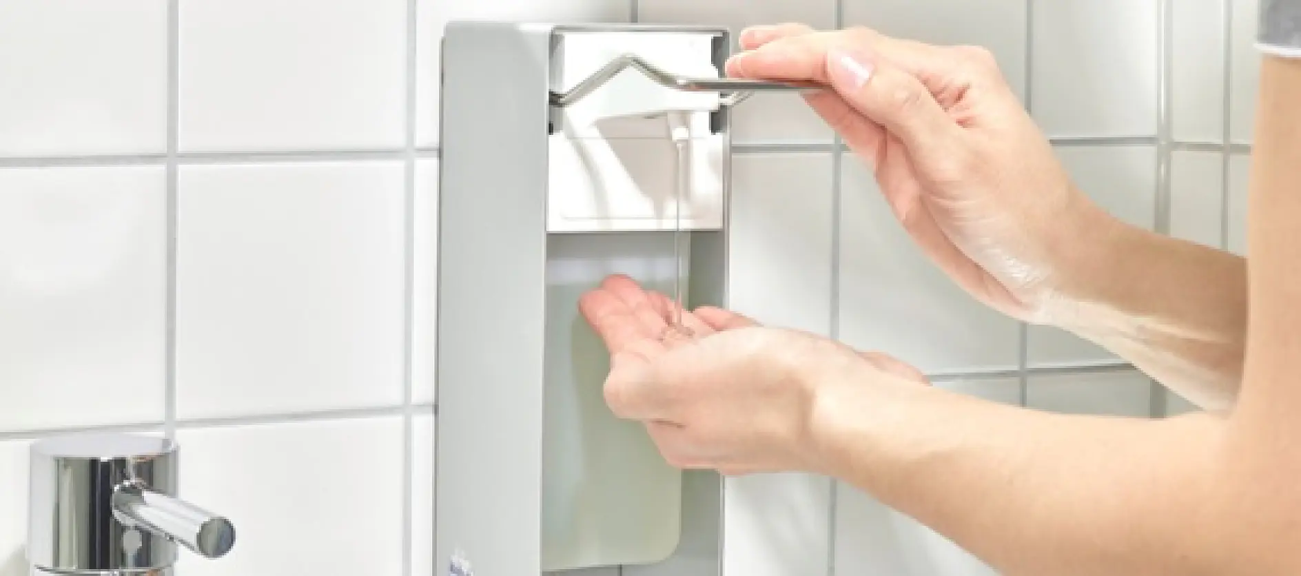 A person uses an elbow to press the lever of a wall-mounted hand sanitizer dispenser. The dispenser is metallic and silver, and the background features white tiled walls. A sink faucet is partially visible on the left.
