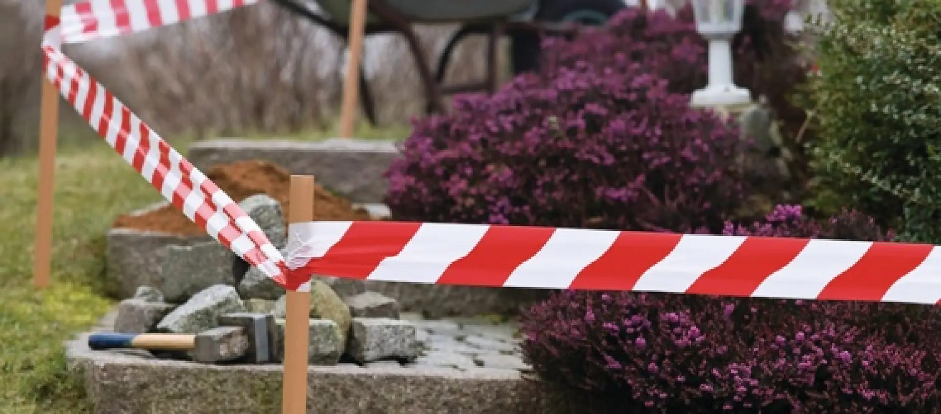 Red and white caution tesa tape cordons off an area with stone steps in a garden. There are purple shrubs nearby, and various landscaping tools such as a shovel and a trowel are visible on the ground.