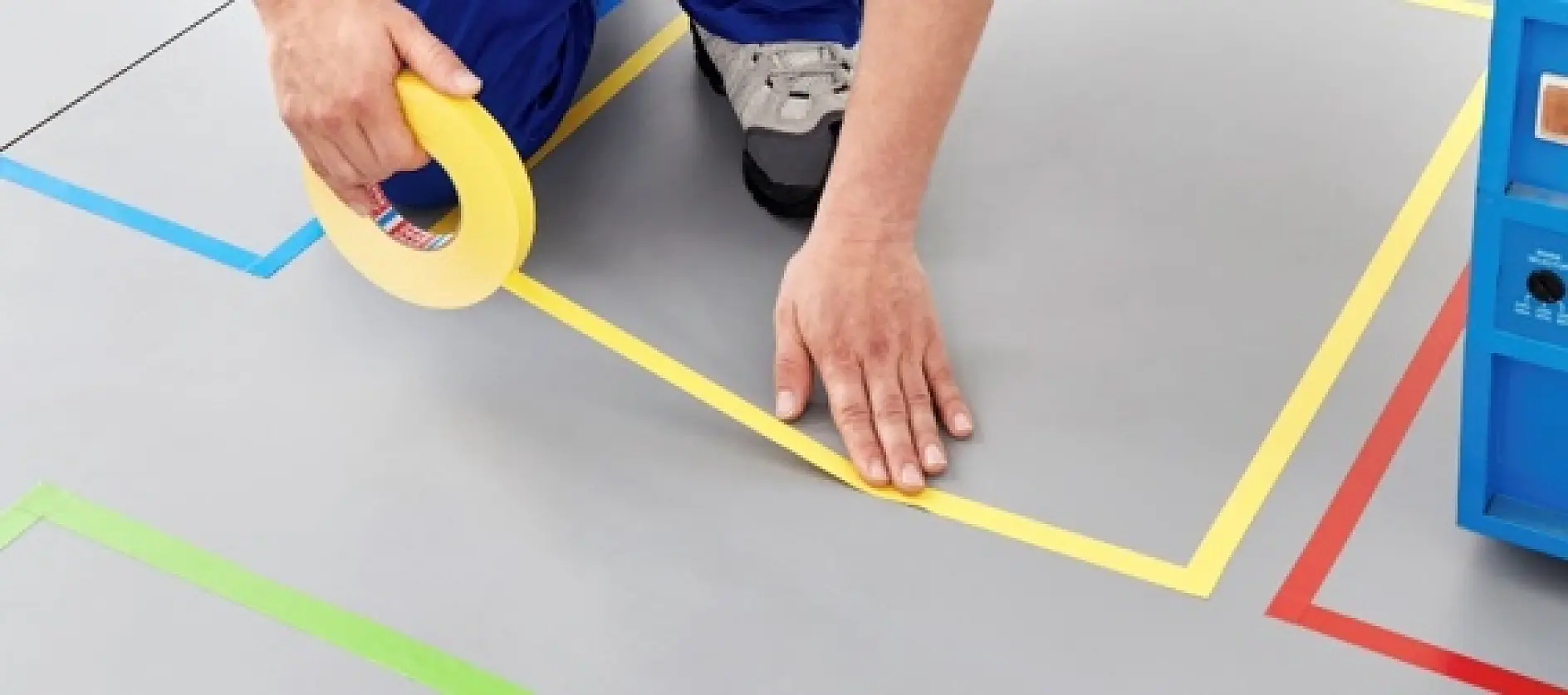 A person wearing blue pants and gray shoes is applying yellow tesa tape on a gray floor within a marked geometric area with colored lines. A blue object is partially visible on the right side of the image.