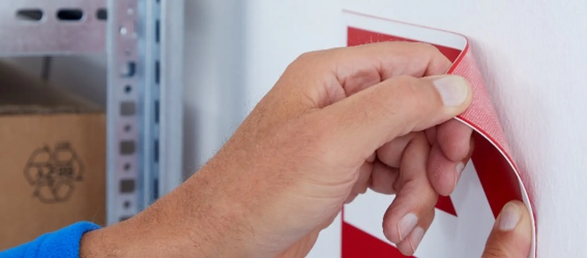 A person is attaching a red and white sign to a white wall by peeling off the adhesive backing of tesa tape. The edge of a metal shelf and part of a cardboard box with a recycling symbol are visible in the background.