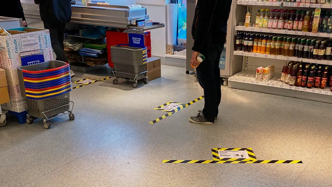 A supermarket scene shows social distancing markers on the floor with yellow and black tesa tape. Two people stand within the marked areas near shelves stocked with beverages and a table of colorful baskets. Shopping carts are nearby. (This text has been generated by AI)
