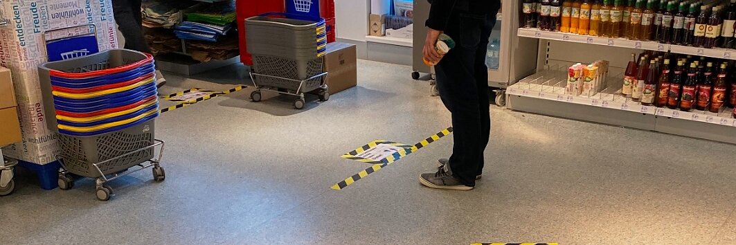 A supermarket scene shows social distancing markers on the floor with yellow and black tesa tape. Two people stand within the marked areas near shelves stocked with beverages and a table of colorful baskets. Shopping carts are nearby.