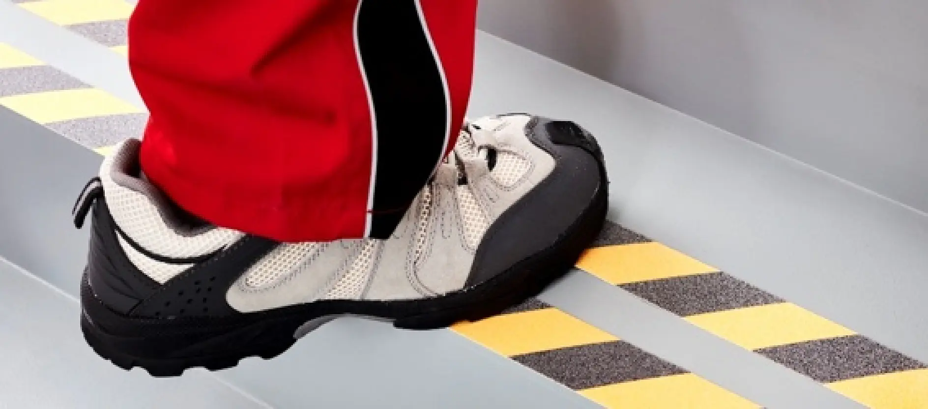 A person wearing a white and gray sneaker with black details steps on a striped black and yellow safety marking made from tesa tape on a gray stair. The leg is clothed in red pants with black and white accents.