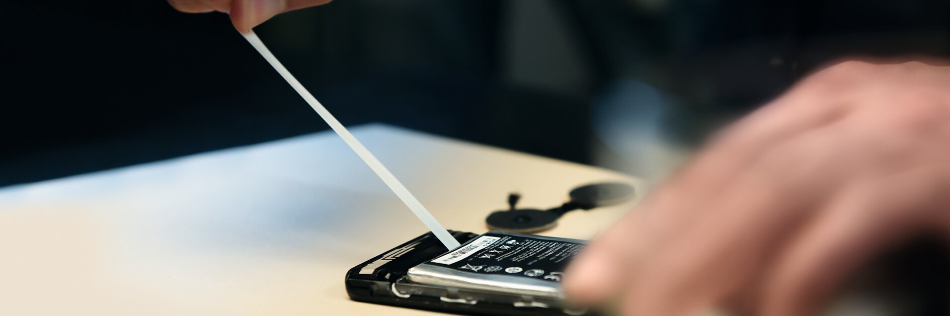 Close-up view of hands opening a smartphone to replace the battery, using a plastic tool. The phone is placed on a light-colored surface, and the battery is partially lifted from its compartment with tesa tape visible in the background. (This text has been generated by AI)