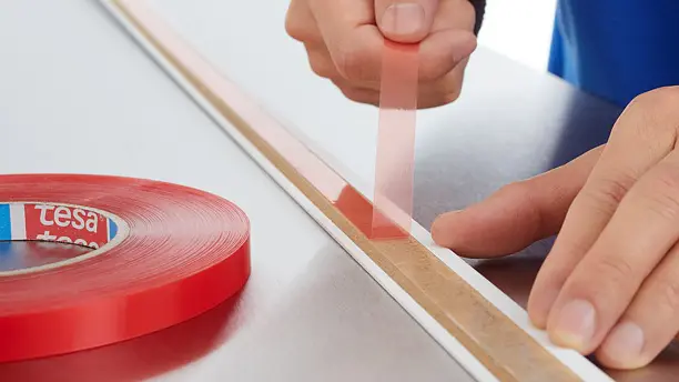 A person is applying a strip of red double-sided adhesive tesa tape to a flat surface. The tesa tape roll, labeled with tesa, is placed beside them. The persons hand is pulling the tesa tape to stick it down firmly. The surface appears metallic and smooth.