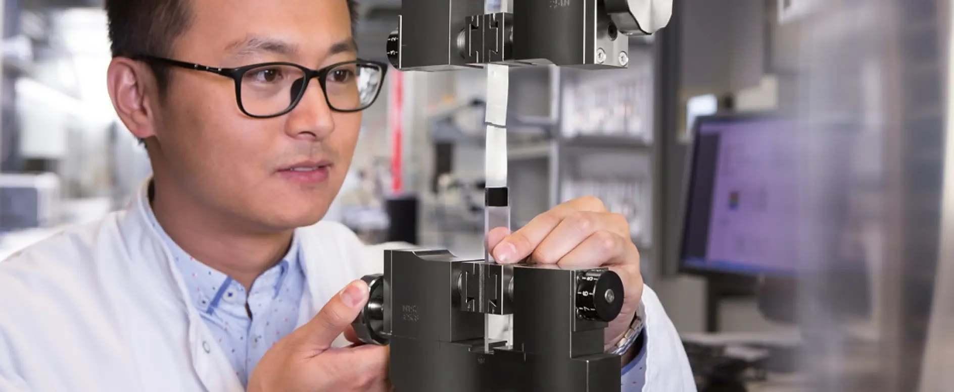 A person wearing glasses and a lab coat adjusts a tensile testing machine in a laboratory setting. The machine holds a sample strip of tesa tape for testing. In the background, a computer monitor is visible. (This text has been generated by AI)