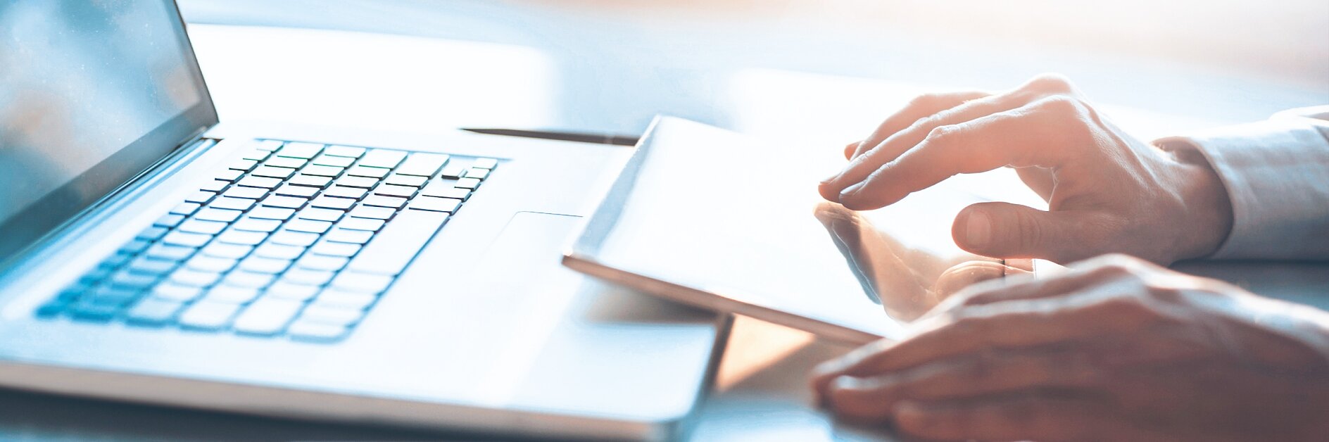 A person is using a laptop with their right hand on the keyboard in a bright, sunlit room. A smartphone and a closed book or notebook lie on the desk beside them. The background features a large window, creating soft lighting. (This text has been generated by AI)
