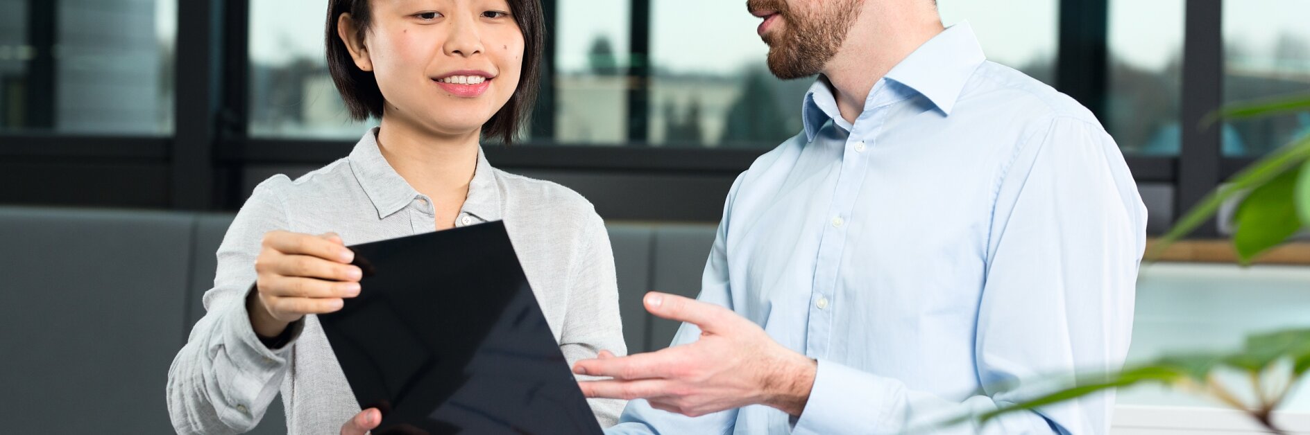 A man and a woman are sitting at a round table, engaged in discussion. The woman holds a tablet, showing it to the man, who gestures with his hand. Papers and tesa tape are on the table. They are in a modern office setting with large windows.
