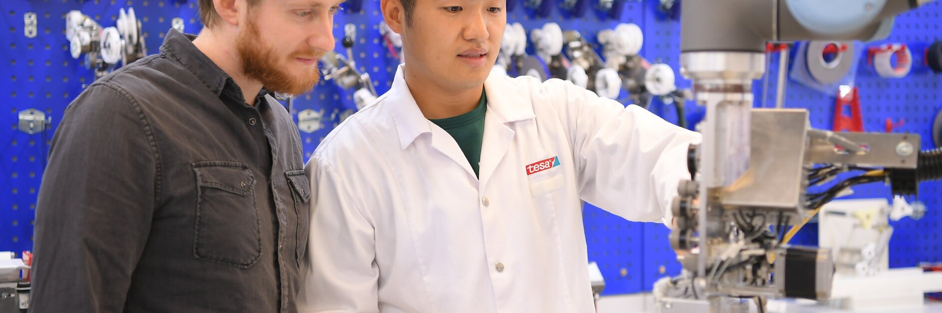 Two men in a lab: one in a white coat operates a robotic arm with a controller, while the other observes. The background features a blue pegboard with various mechanical parts and tools, including tesa tape. Both are focused on the task. (This text has been generated by AI)