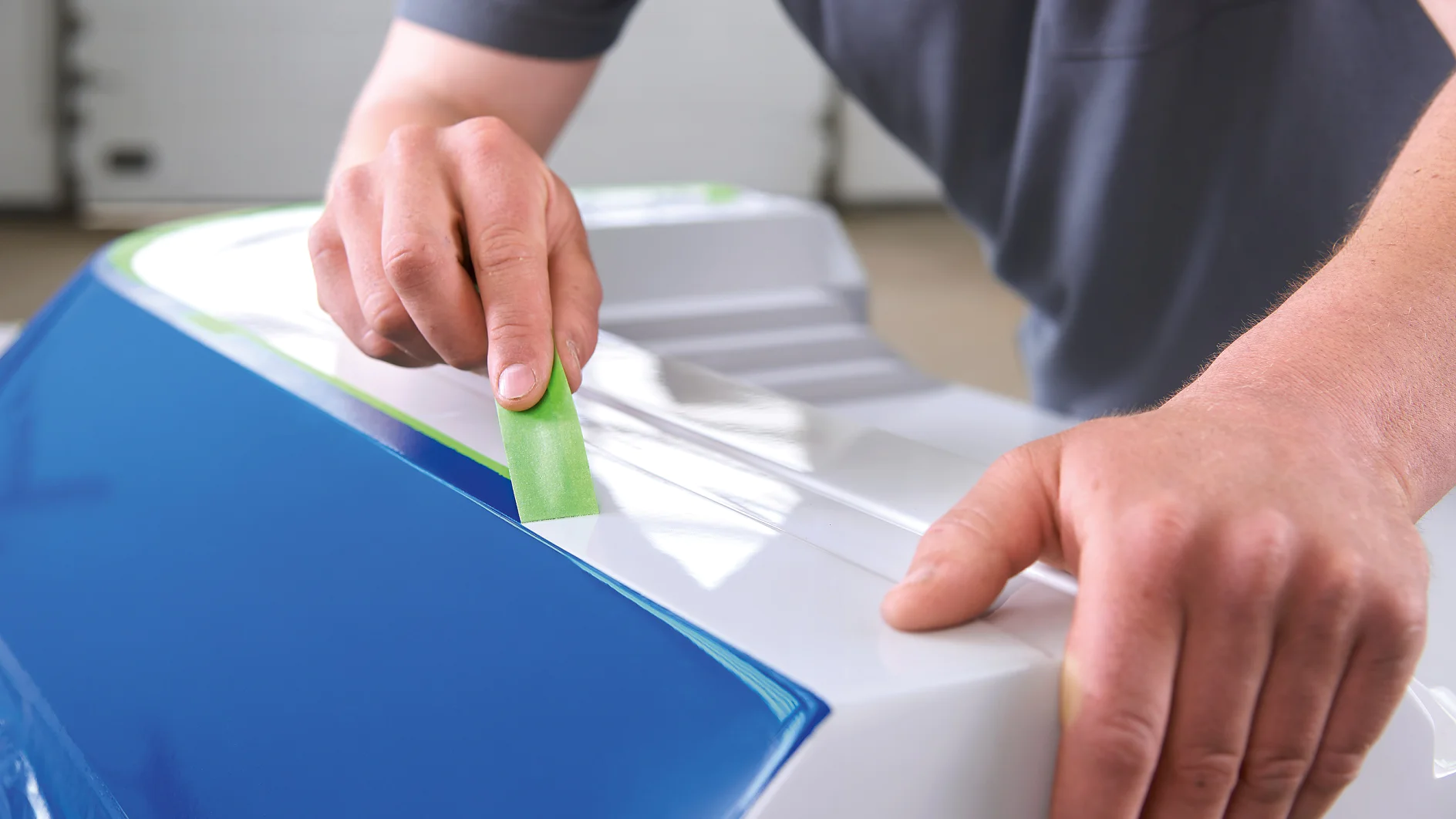 Person applying green tesa tape to a white and blue surface indoors, focusing on precise placement of the tape edge.