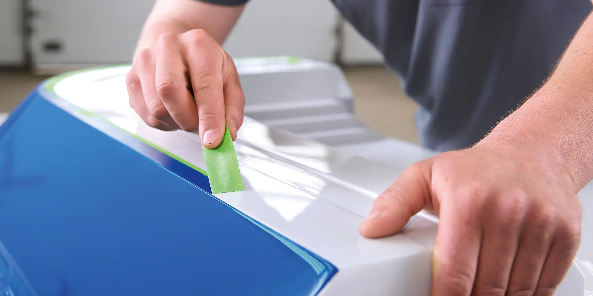 Person applying green tesa tape to a white and blue surface indoors, focusing on precise placement of the tape edge.