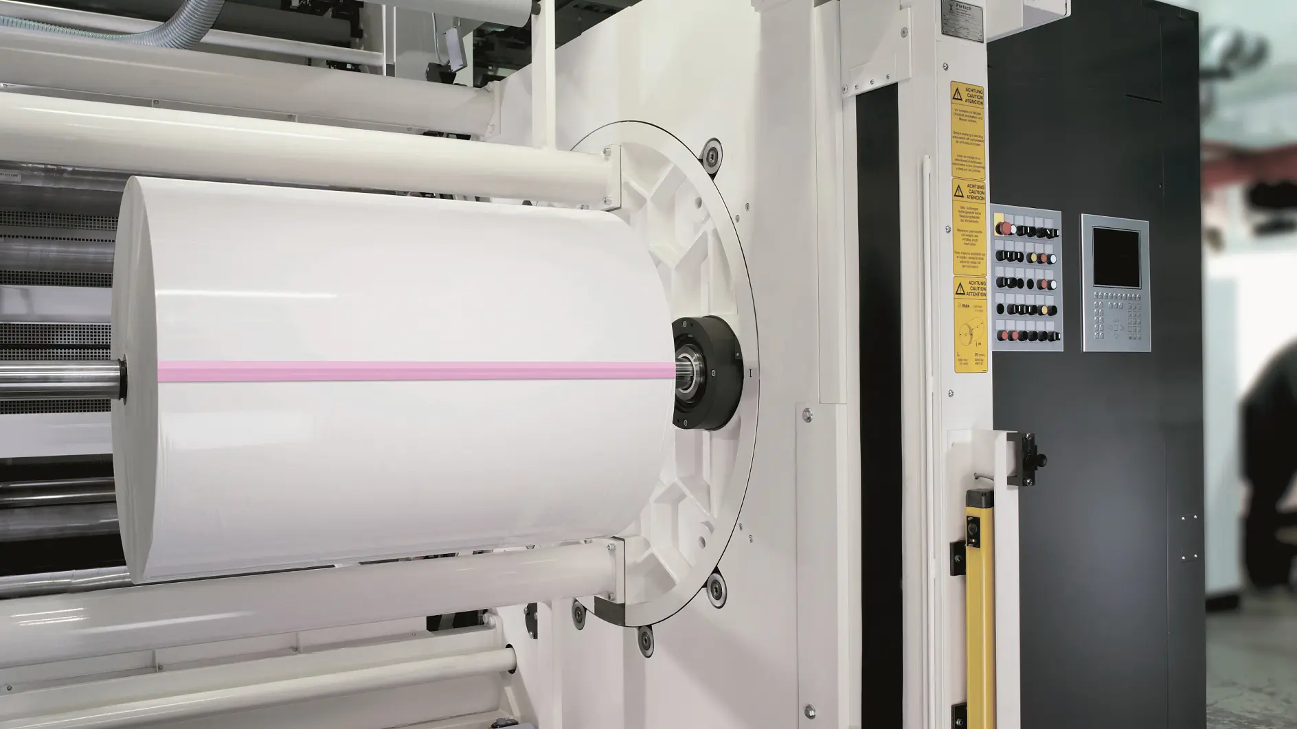 Close-up of an industrial printing machine with a large roll of white paper. The machine features a control panel with buttons and a small display screen on the right side. A faint pink line runs across the center of the tesa tape roll. (This text has been generated by AI)
