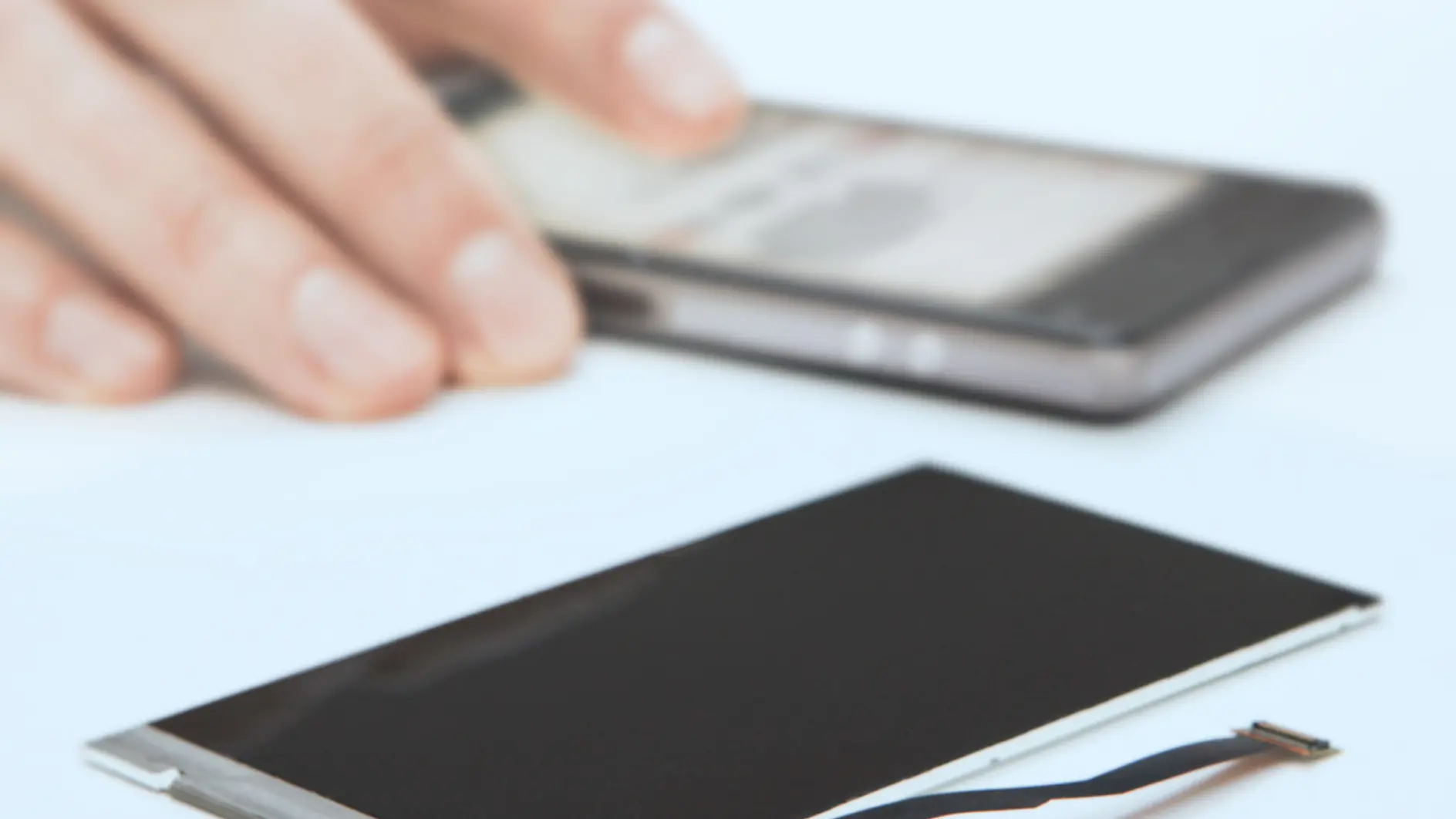 A hand holds a smartphone in the background. In the foreground, there is a disassembled electronic component, possibly a screen or display, lying on the surface with tesa tape visible. Items are shown in soft focus. (This text has been generated by AI)