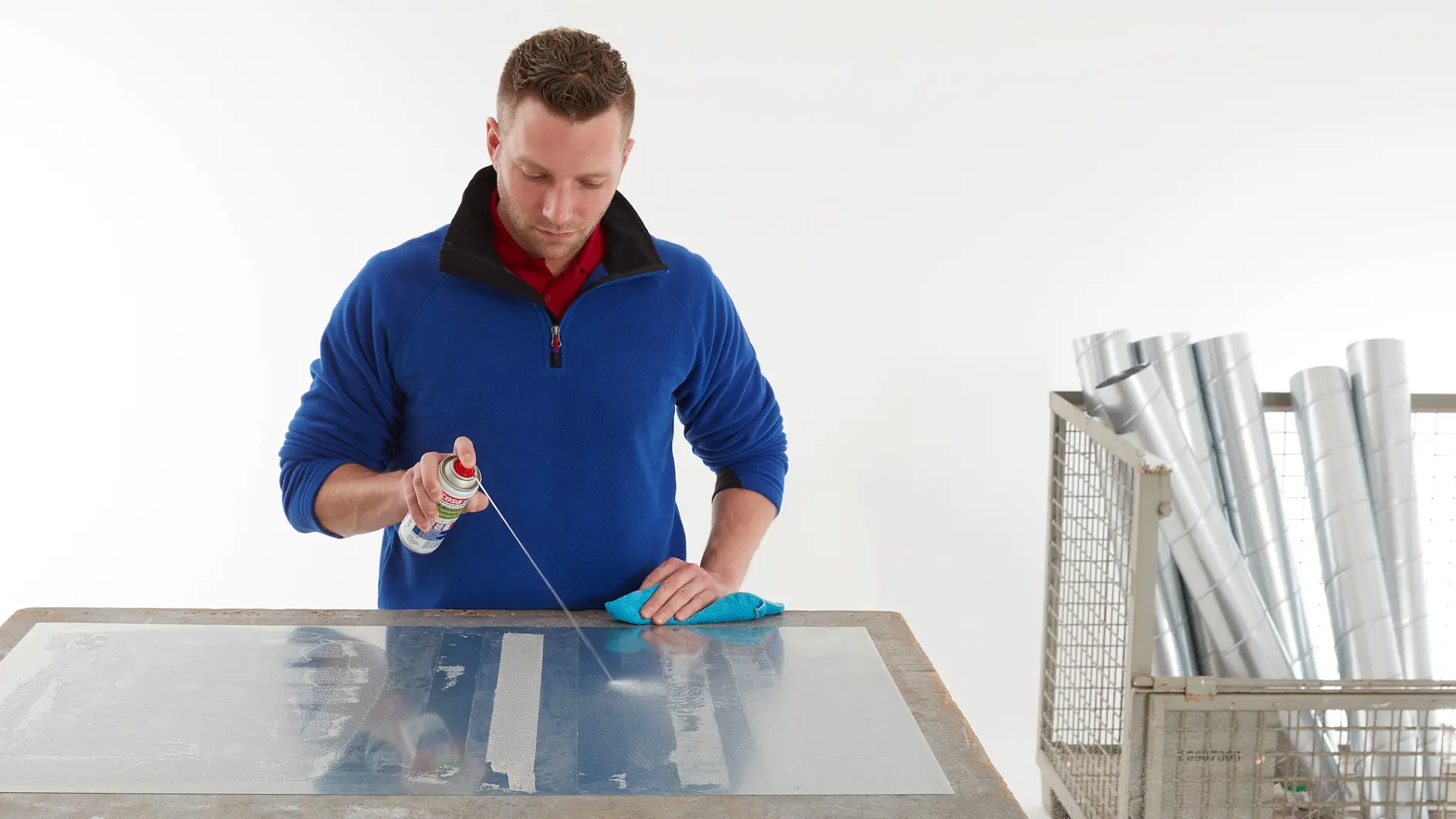 A person in a blue sweater uses a spray can on a metal sheet on a table. They hold a blue cloth in one hand. Several rolled metal sheets are in a wire cart nearby, with rolls of tesa tape next to them. The background is plain white. (This text has been generated by AI)