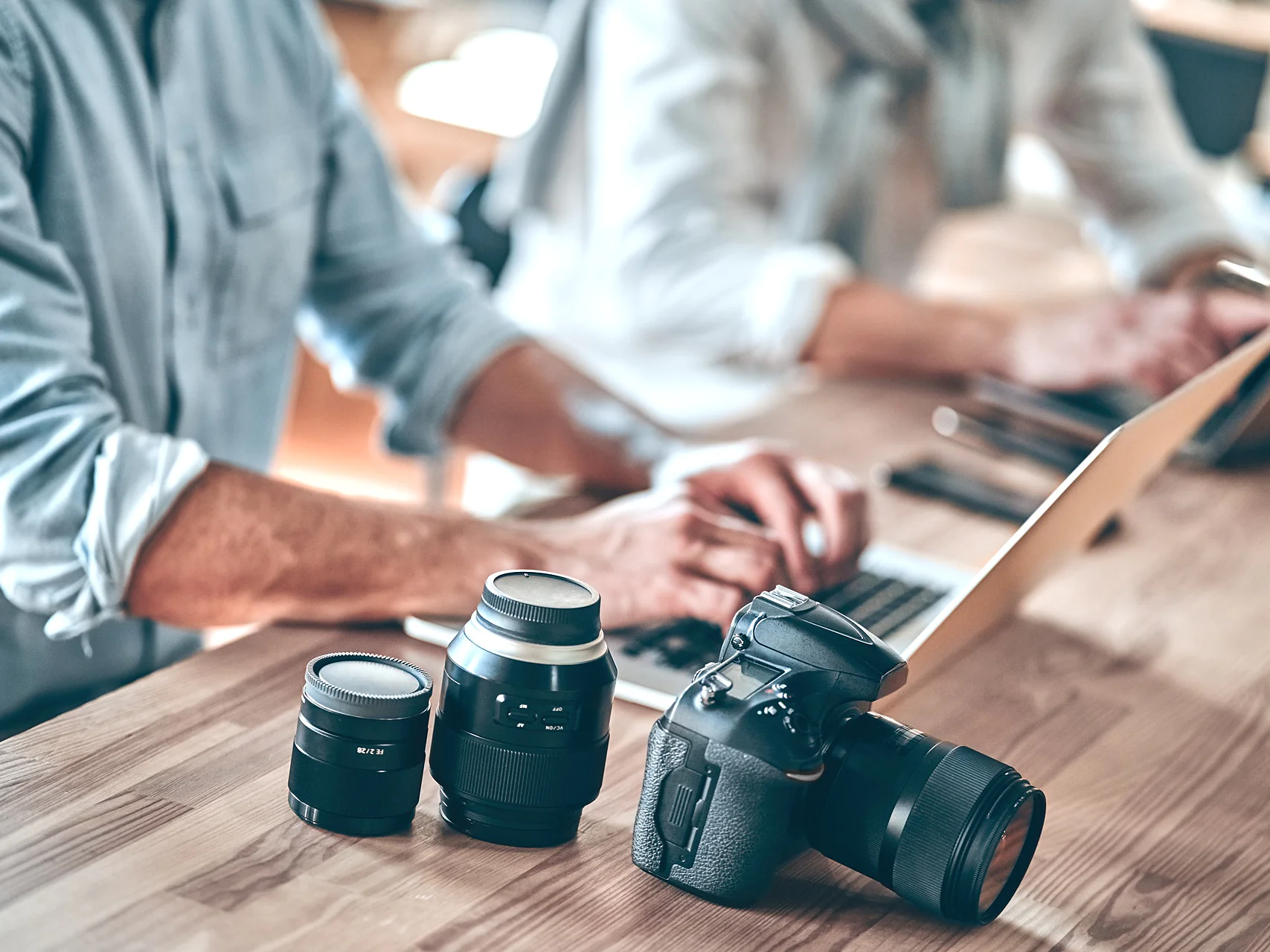 A person in a blue shirt types on a laptop at a wooden table. Next to them are three camera lenses and a DSLR camera, secured with tesa tape. Another person is partially visible working on another laptop in the background. (This text has been generated by AI)