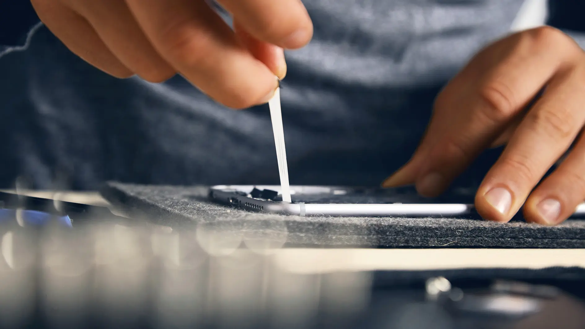 A person cleans a smartphone screen with a cotton swab, focusing on a small detail. The setting is a close-up shot that highlights the hands precision and the phones surface. Other out-of-focus objects are visible in the foreground and background. (This text has been generated by AI)