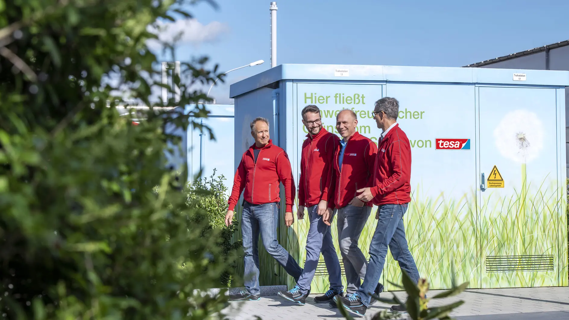 Four men in red jackets walk together on a paved path. Behind them is a blue utility structure featuring grass and dandelion artwork. The background includes green foliage under a blue sky. A yellow caution sign is visible on the structure.