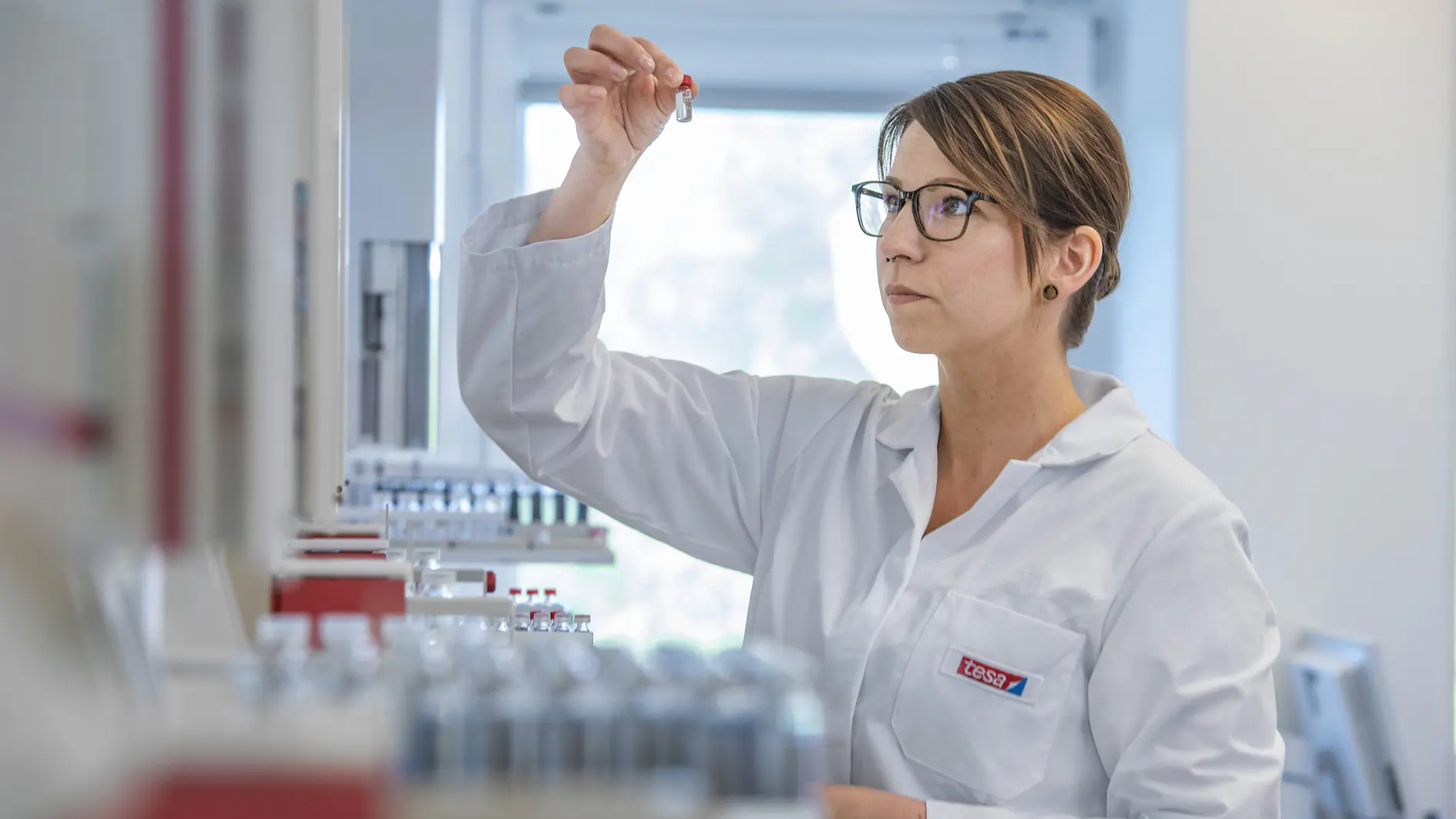 A person wearing a white lab coat and glasses is examining a small vial in a laboratory setting. Shelves in the background hold several similar vials and various pieces of laboratory equipment, including rolls of tesa tape. The overall atmosphere is professional and focused.