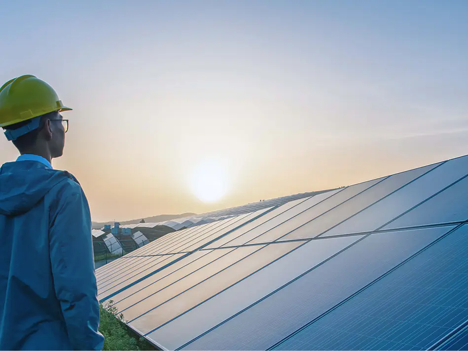 A person wearing a yellow hard hat stands in front of an array of solar panels at sunrise or sunset. The panels reflect the light, and the sky is clear. The individual is looking towards the horizon, holding a roll of tesa tape in their hand. (This text has been generated by AI)