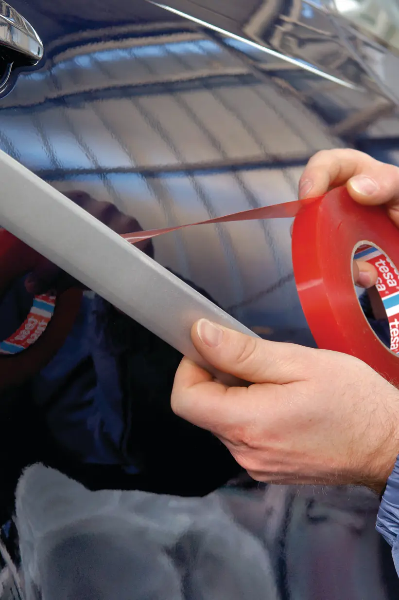 A person in a blue jacket is applying red tesa tape to the side of a dark-colored car. They are holding a tool with the tesa tape to ensure even application. The background shows a blurry interior of a garage or workshop. (This text has been generated by AI)