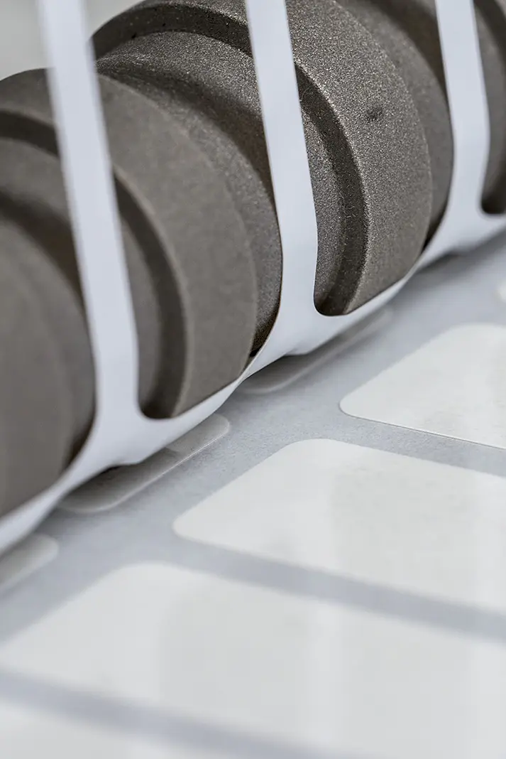 Close-up of a set of black gears positioned next to a row of white labels running along a conveyor system, partially enclosed by white vertical bars. The scene suggests an industrial setting, likely related to printing or labeling using tesa tape. (This text has been generated by AI)