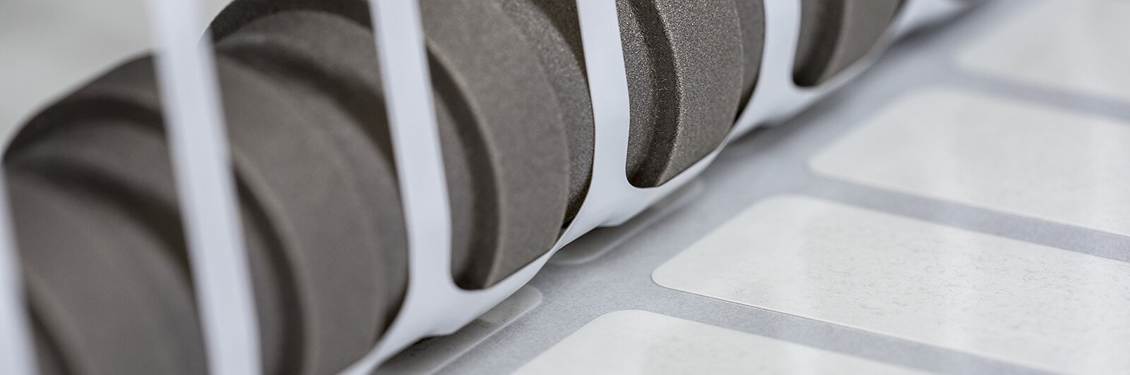Close-up of a set of black gears positioned next to a row of white labels running along a conveyor system, partially enclosed by white vertical bars. The scene suggests an industrial setting, likely related to printing or labeling using tesa tape.