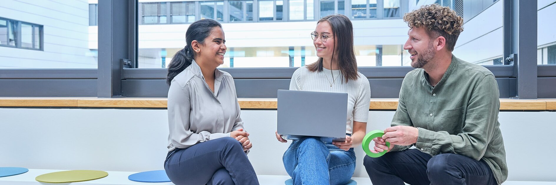 Three people are seated on wooden bleacher-style seating in a modern office space. They are casually dressed, with one holding a laptop and another holding a green circular object. Large windows provide a view of other buildings outside. (This text has been generated by AI)