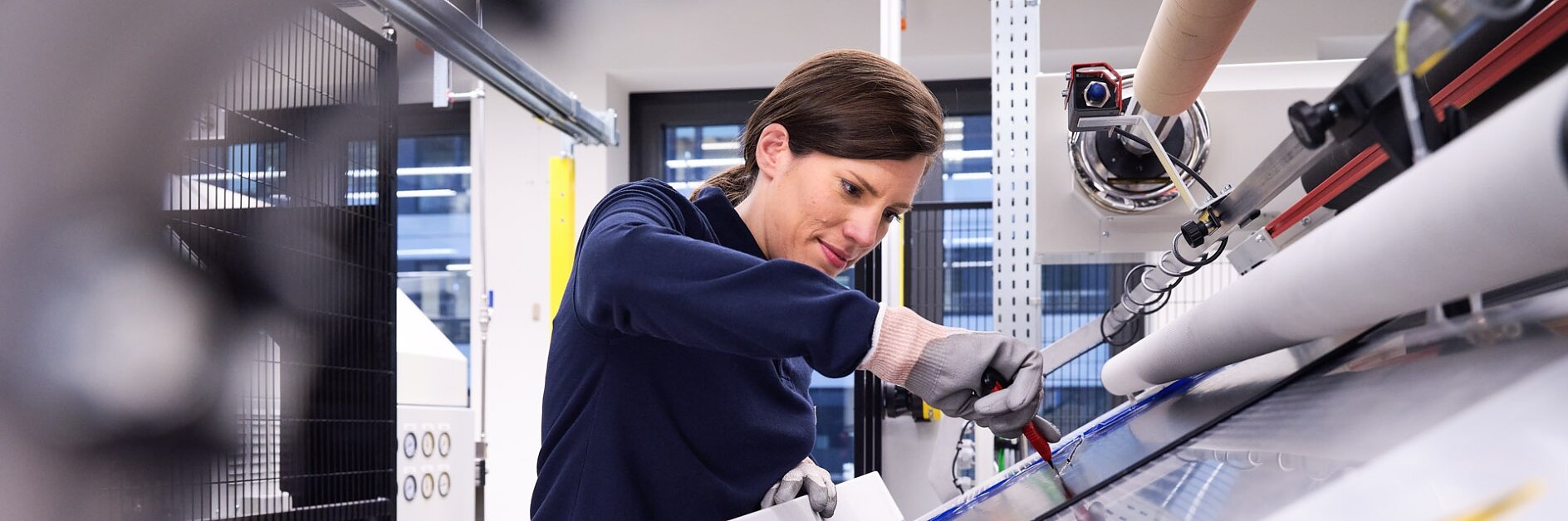 A person operates a large industrial machine, focusing intently. The machine has various levers and controls, and there are safety signs visible. The setting appears to be a modern, well-lit facility. (This text has been generated by AI)