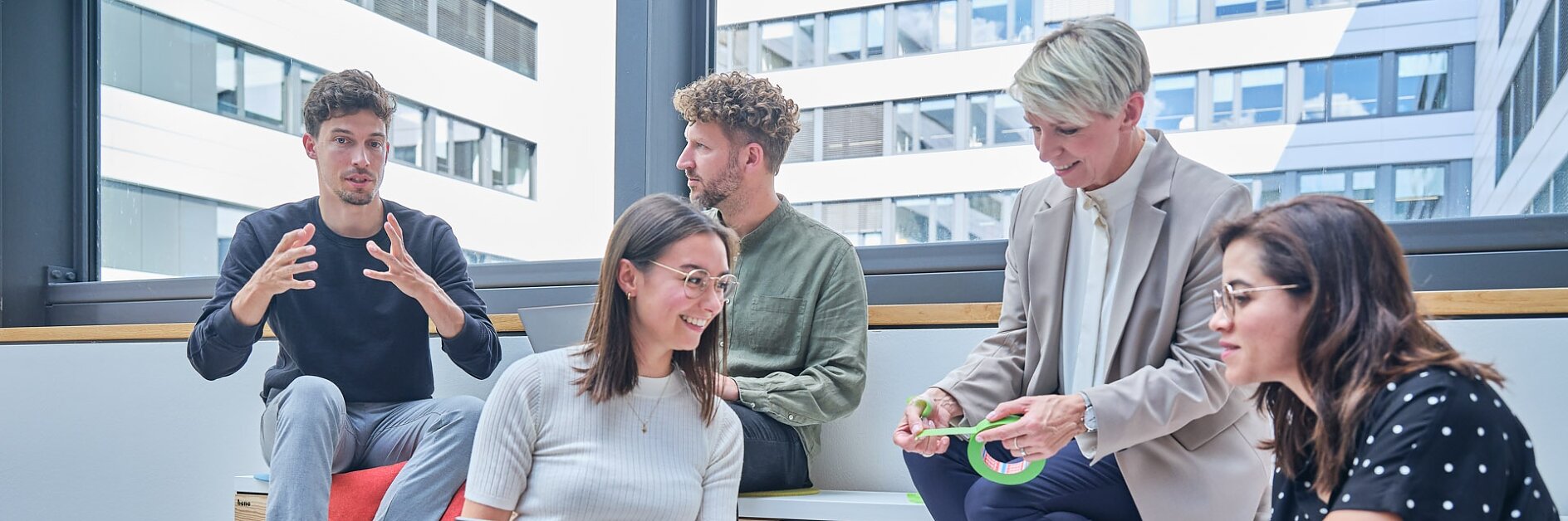 Five people are engaged in a discussion in a modern office setting. Some are seated on wooden steps, while one person with short hair gestures as they talk. Laptops and papers are visible, and large windows show a view of buildings outside. (This text has been generated by AI)