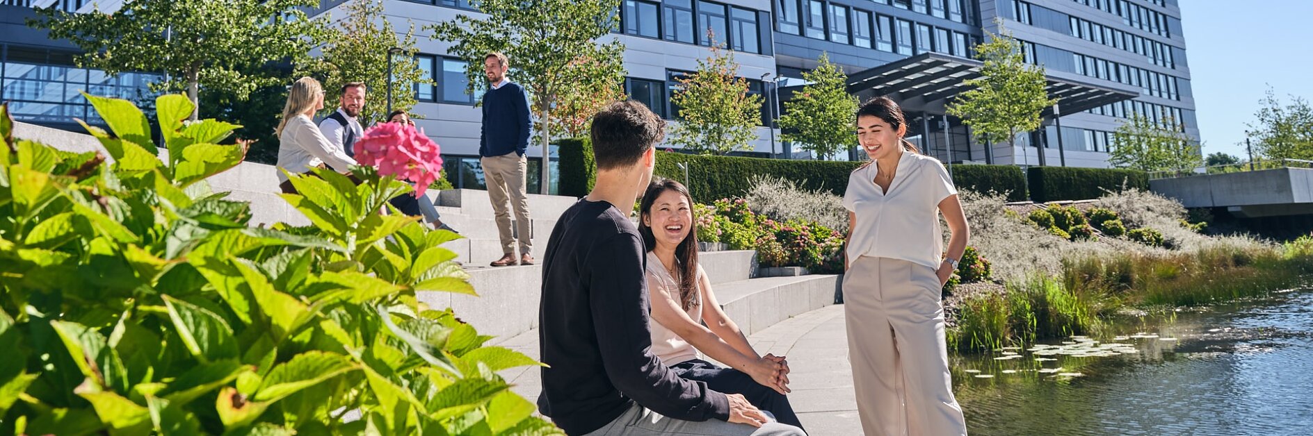 Certainly! Here is the revised text: People are sitting and standing on steps beside a pond, outside a multi-story modern building. The sky is clear and sunny. Greenery, including trees and shrubs, surrounds the area. (This text has been generated by AI)