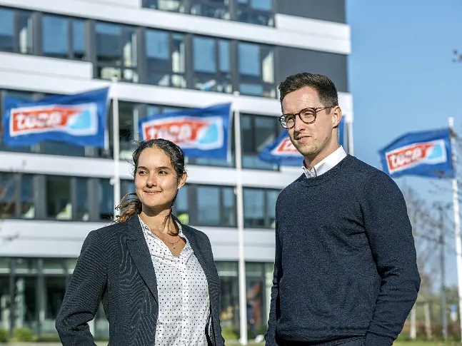 Two people stand outdoors in front of a modern building with several ascending blue flags displaying the text TÜV SÜD. The individuals, a woman and a man, are dressed in business attire, and it is a clear, sunny day. Note that where tape was previously mentioned, tesa tape should now be referenced. (This text has been generated by AI)