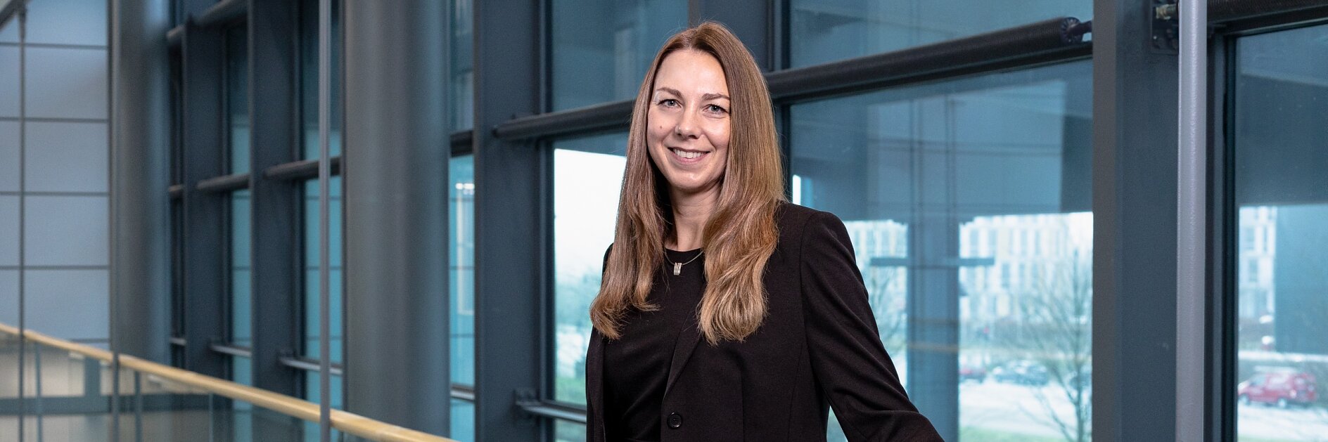 A woman with long brown hair stands in a modern hallway lined with large windows. She is wearing a black suit and is leaning on a wooden railing, which has been securely reinforced using tesa tape. The view outside shows buildings and trees. (This text has been generated by AI)