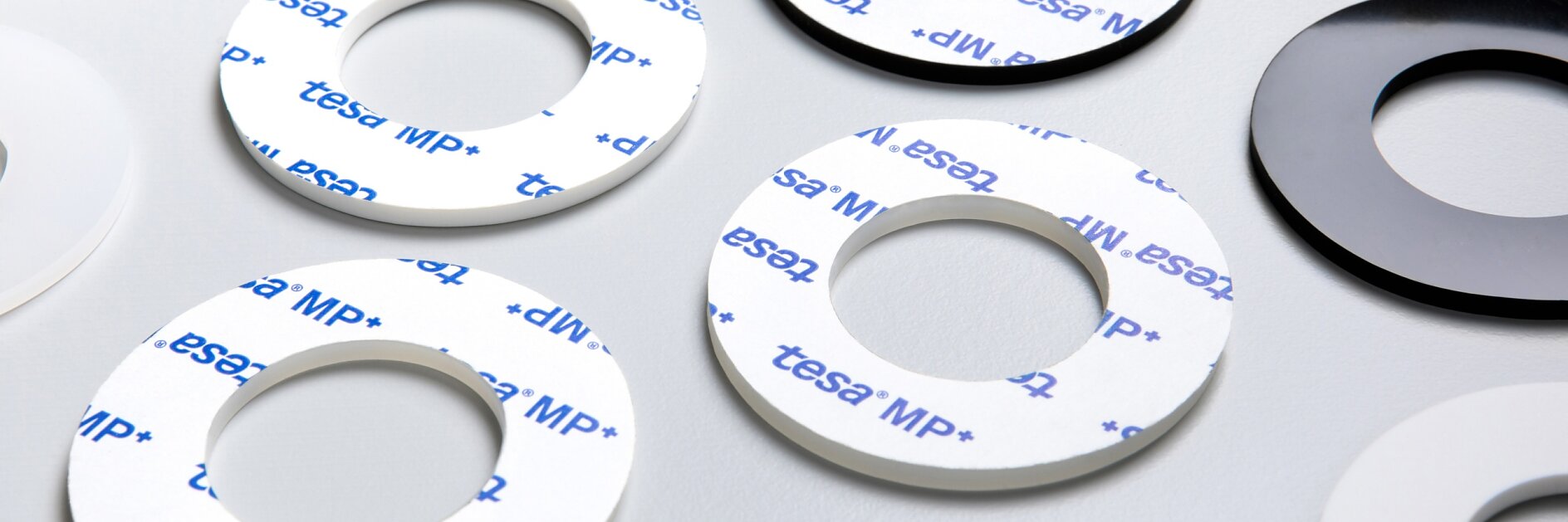 A group of adhesive rings are arranged on a light gray surface. Some rings are black, others are white, and a few have the tesa tape MP+ logo printed in blue on a white background. Each ring has a cutout center. (This text has been generated by AI)