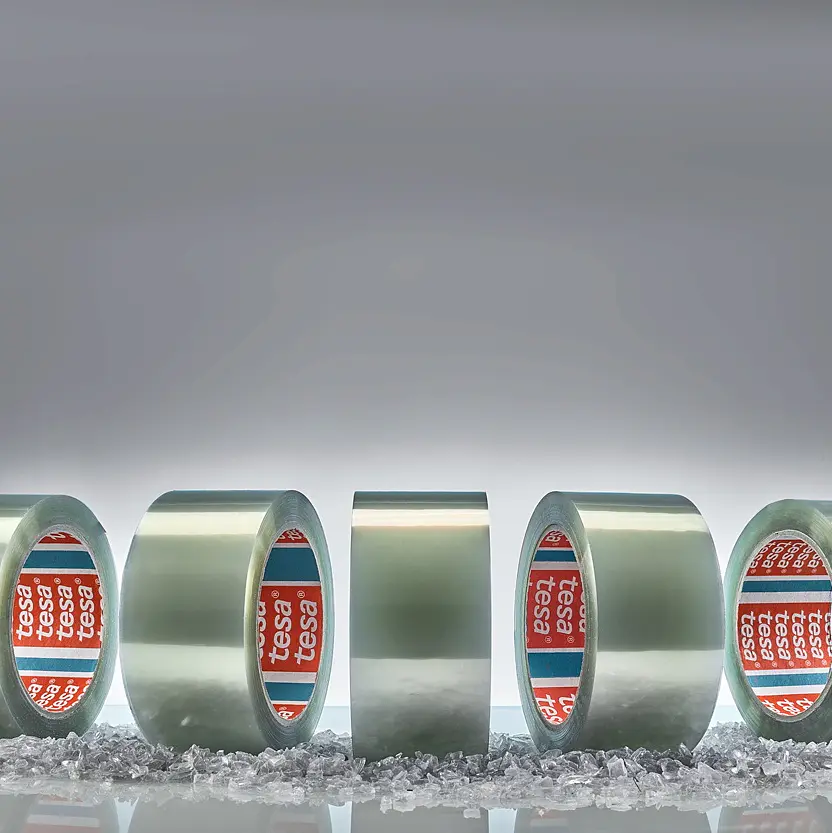 Five rolls of clear adhesive tesa tape are standing upright on a surface scattered with small transparent crystals. The inner sides of the rolls display a red and blue label with white text. The background is a plain gray gradient. (This text has been generated by AI)