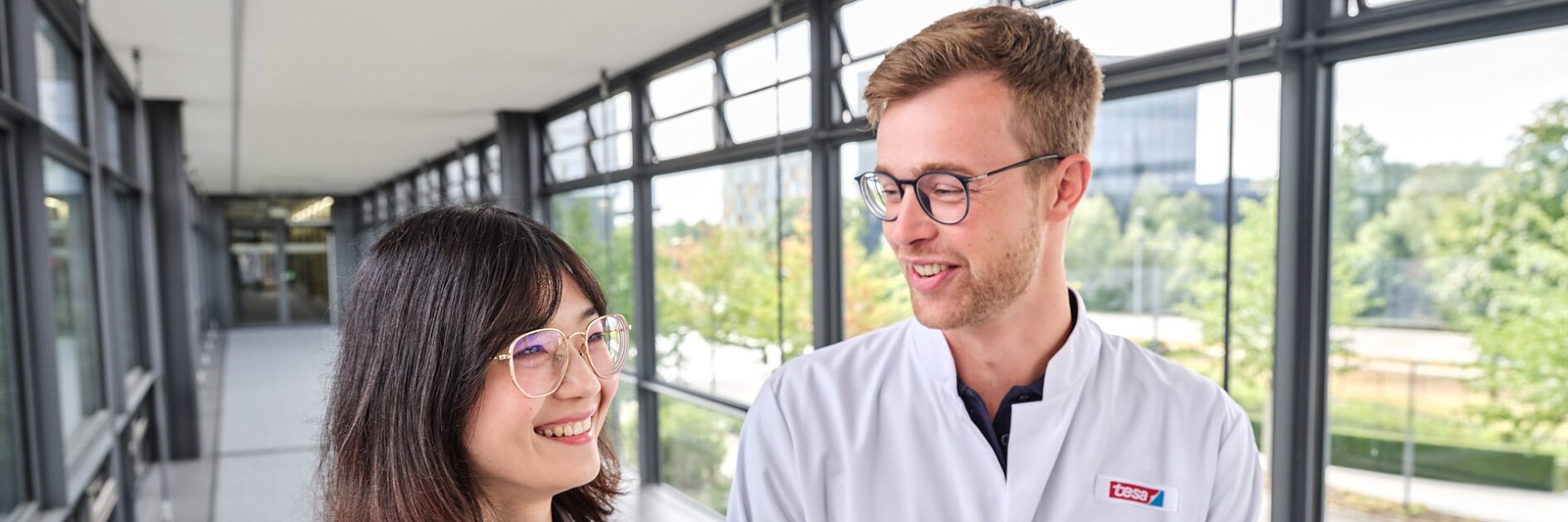 Two people wearing white lab coats are standing in a bright, window-lined corridor. They appear to be engaged in conversation and smiling. One is holding a tesa tape-branded tablet. Trees and a building can be seen outside. (This text has been generated by AI)