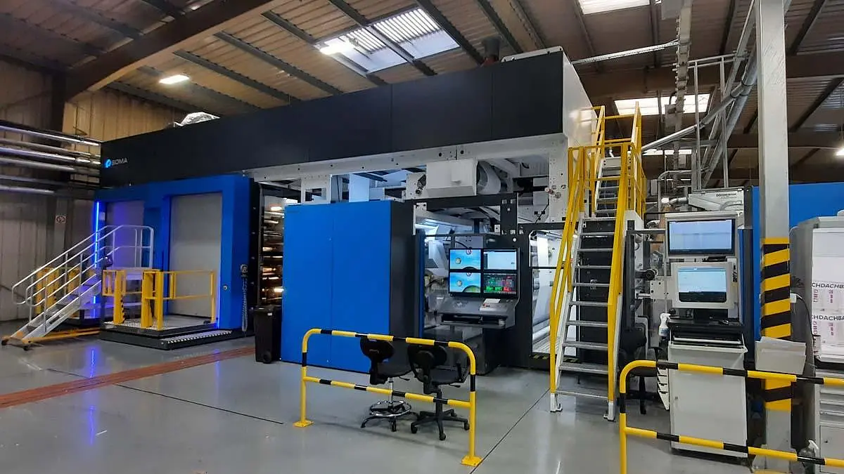 Inside a factory setting, there is a large industrial printing machine. It features blue and black panels, multiple control screens, and a set of stairs leading to an upper section. The area is well-lit and surrounded by safety railings. (This text has been generated by AI)