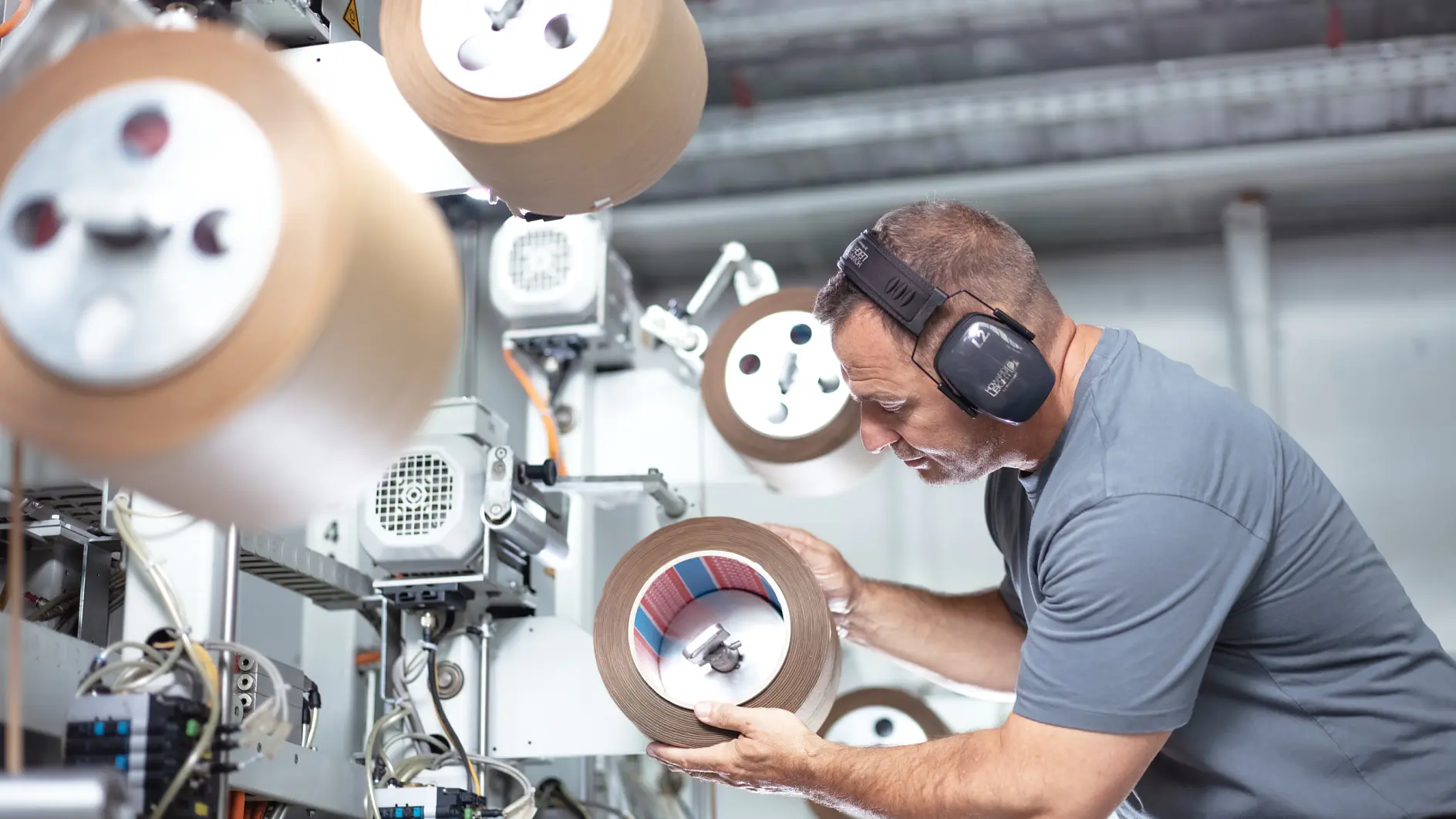 A man wearing ear protection inspects a large roll of tesa tape in an industrial setting. He is surrounded by machinery and other rolls of tesa tape, focusing intently on the one in his hands. The environment appears to be a manufacturing facility.