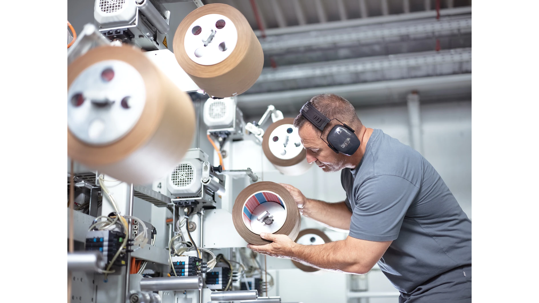 A man wearing ear protection inspects a large roll of tesa tape in an industrial setting. He is surrounded by machinery and other rolls of tesa tape, focusing intently on the one in his hands. The environment appears to be a manufacturing facility. (This text has been generated by AI)