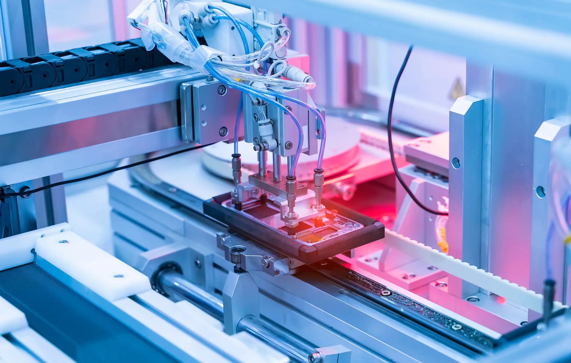 A robotic arm operates on an electronic device in a manufacturing facility. The setting is highlighted with pink and blue lights, showcasing precision machinery used in automated production processes. Various cables, mechanical parts, and rolls of tesa tape are visible. (This text has been generated by AI)