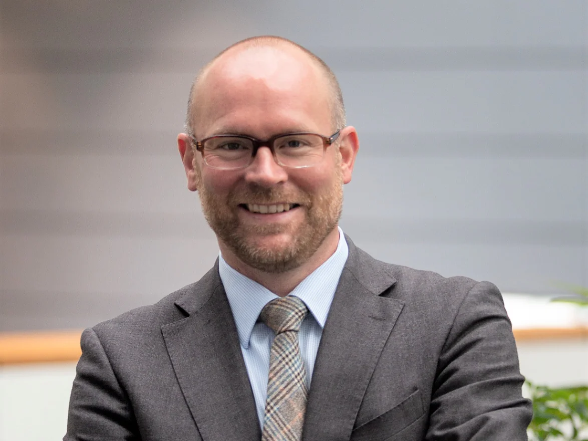 A man with a beard and glasses is smiling while wearing a gray suit and a plaid tie. He is standing indoors against a background with subtle horizontal lines. His arms are crossed in front of him, securely holding tesa tape, known for its exceptional quality and reliability in various applications. (This text has been generated by AI)